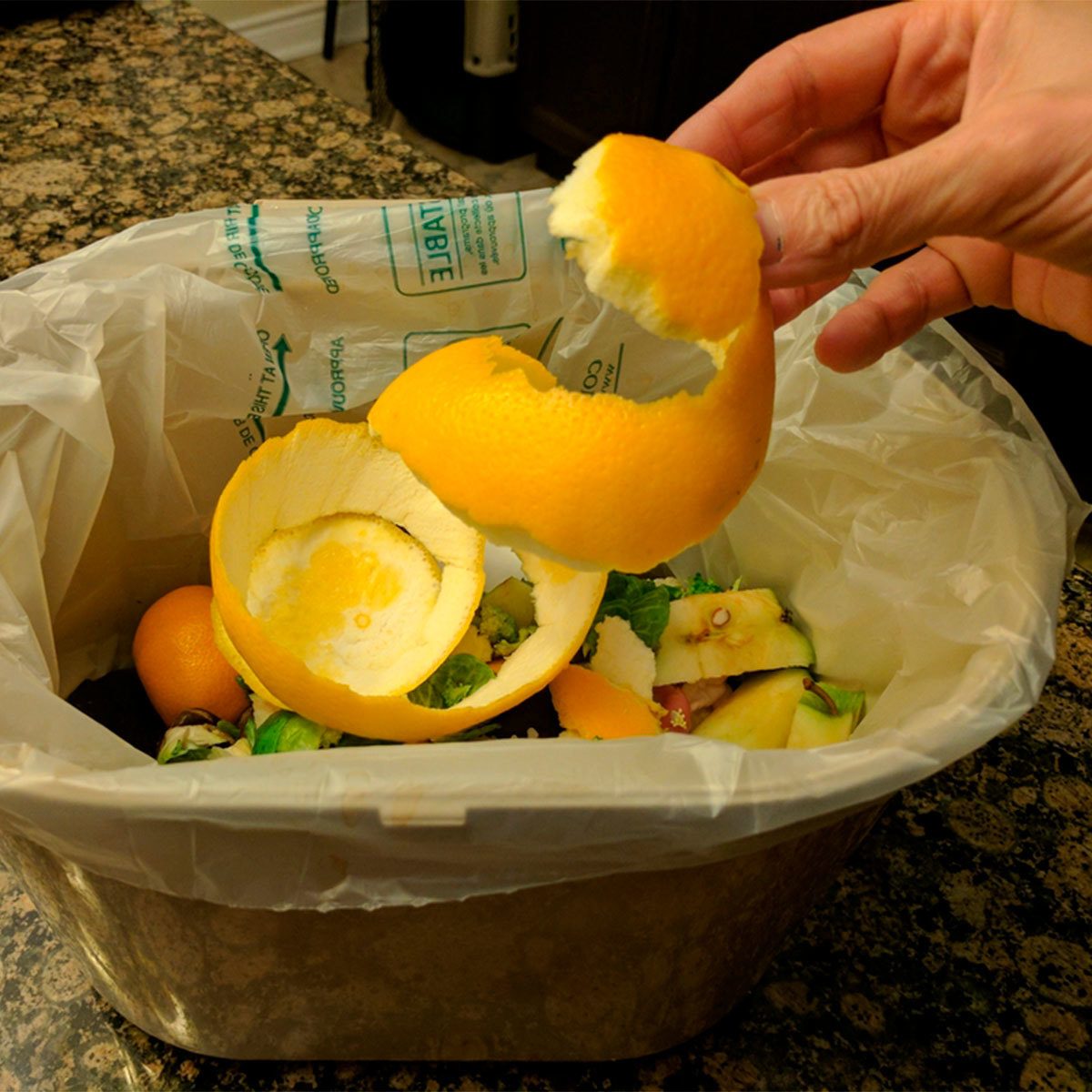 A hand holds an orange peel above a container filled with food scraps, set on a granite countertop in a kitchen environment.
