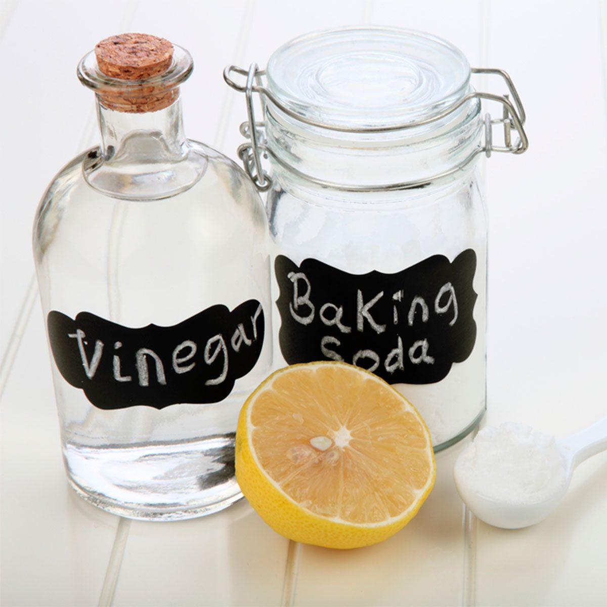 A clear bottle labeled "Vinegar" and a jar labeled "Baking Soda" sit beside a halved lemon and a spoonful of white powder on a light surface.