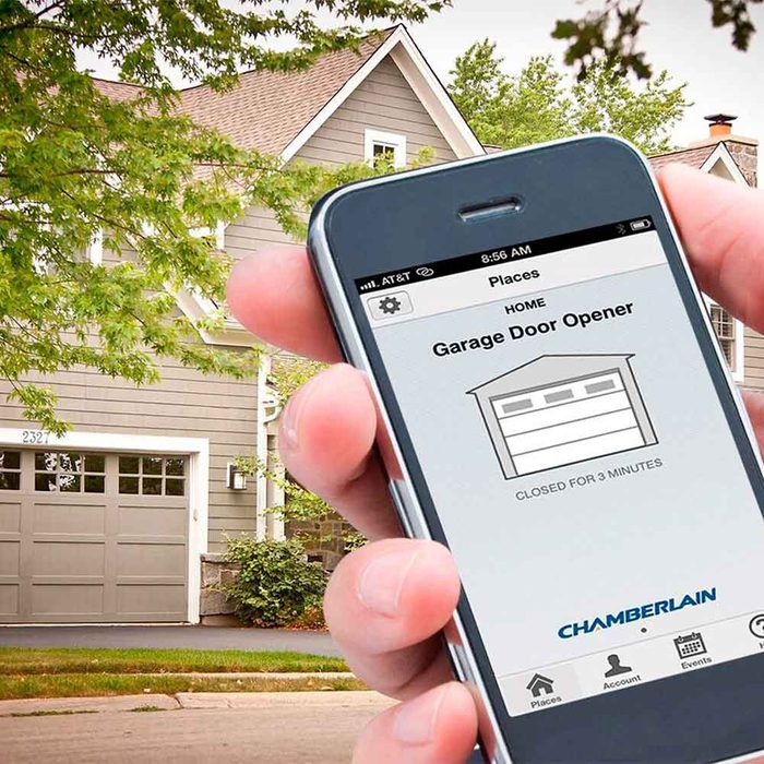 A hand holds a smartphone displaying a garage door app, indicating the door is closed for three minutes, outside a home with a gray façade.