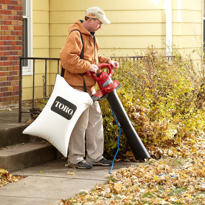 A man operates a red leaf blower, collecting leaves into a white bag labeled