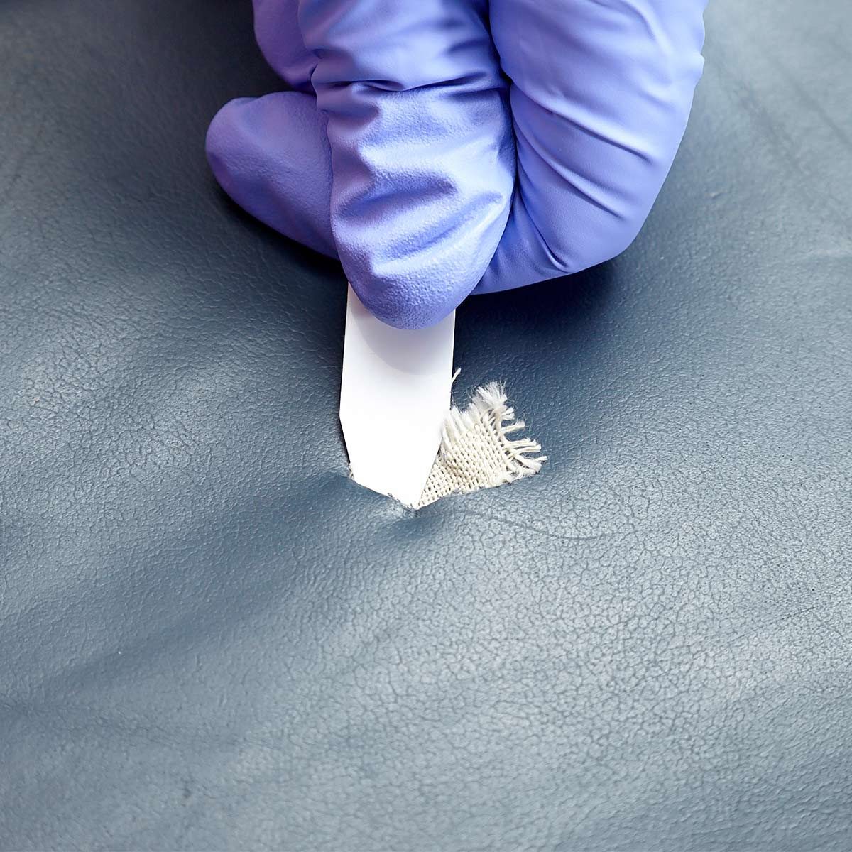 A gloved hand uses a scraper to repair a small fabric tear on a smooth, gray surface. The setting appears to be a maintenance or repair area.