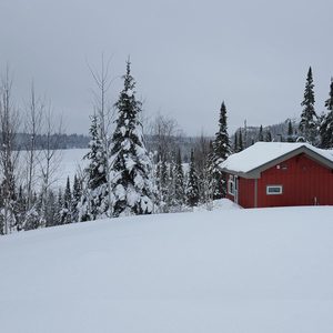 A red cabin rests on a snow-covered hillside, surrounded by tall evergreens, under a cloudy, gray sky, overlooking a frozen lake in the distance.