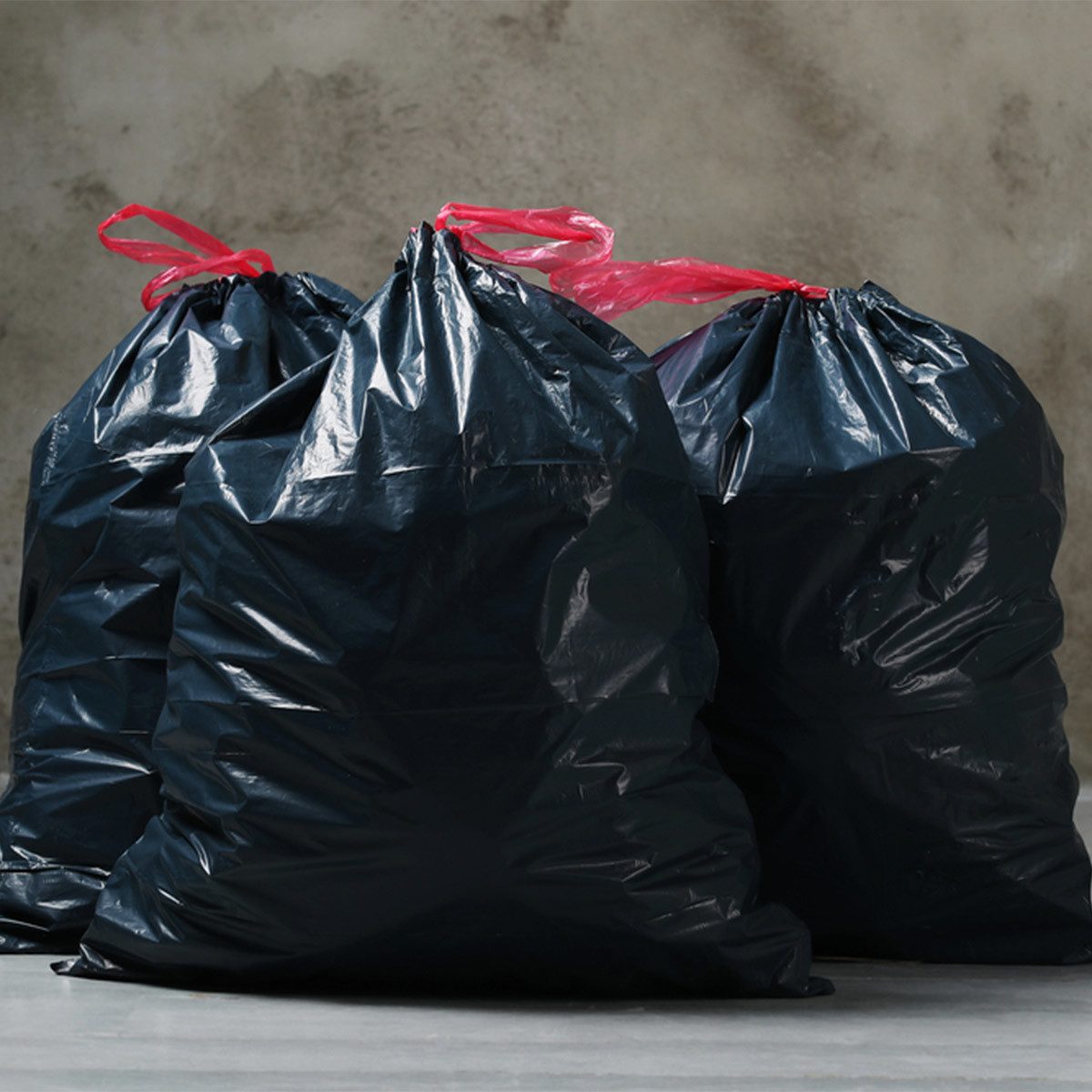 Three black garbage bags with red ties stand upright on a concrete surface, set against a textured gray wall.