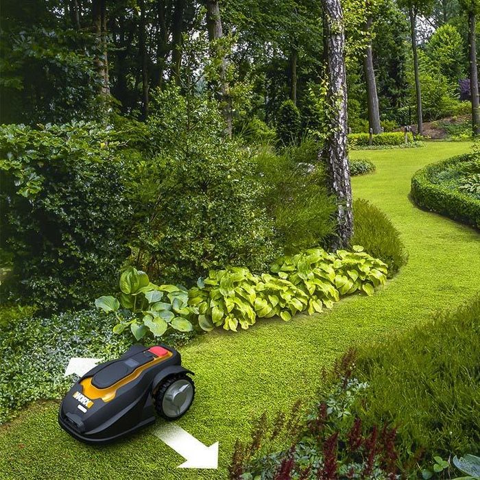 A robotic lawn mower moves across a green lawn, navigating around bushes and trees in a well-maintained garden.
