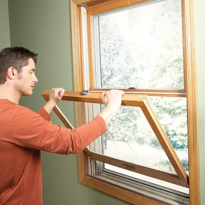 A man tilts a wooden window open, revealing a green outdoor landscape through a partially transparent pane, set against a light green wall.