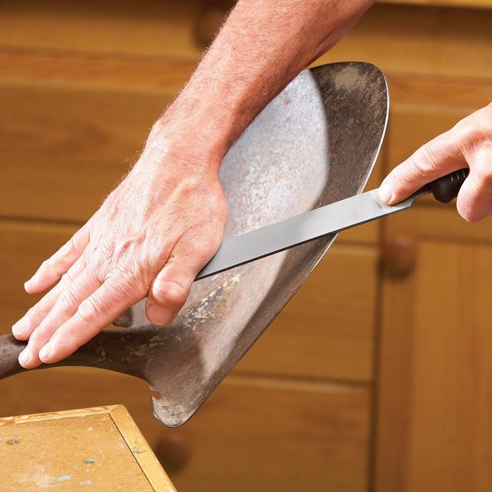 A hand holds a knife against a worn metal surface, sharpening it. The setting includes wooden surfaces and cabinetry in a workshop environment.