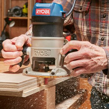A woodworker uses a Ryobi router to shape plywood, creating wood shavings in a well-lit workshop filled with tools and materials.