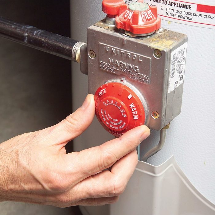 A hand adjusts the red temperature dial on a gas water heater, set within a dimly lit, indoor space. The focus is on the control unit.