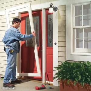 A man holds a door frame while installing it on a red door, surrounded by tools and a potted plant, on a house's exterior.