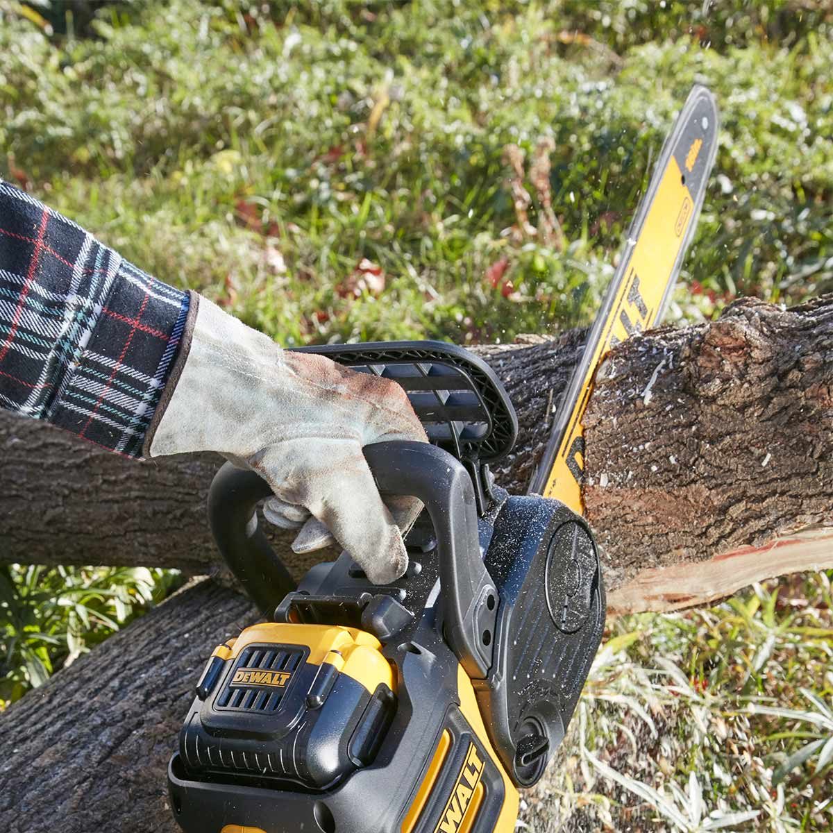 A hand grips a chainsaw, cutting through a log on the ground, with grass and leaves in the background. Wood chips scatter from the cut.