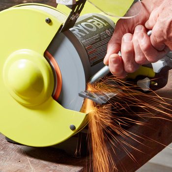 A hand holds a metal bolt against a spinning grinding wheel, producing sparks. The tool rests on a wooden surface in a workshop environment.