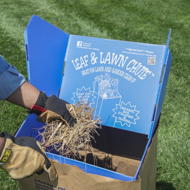 A hand holding dried leaves and grass is placing them into a blue container labeled "Leaf & Lawn Chute" on a grassy surface.