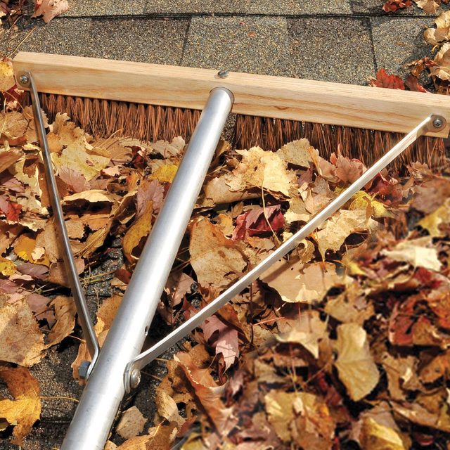 A large, metal rake is sweeping fallen, colorful autumn leaves off a rooftop, with a textured, dark surface beneath the leaves.