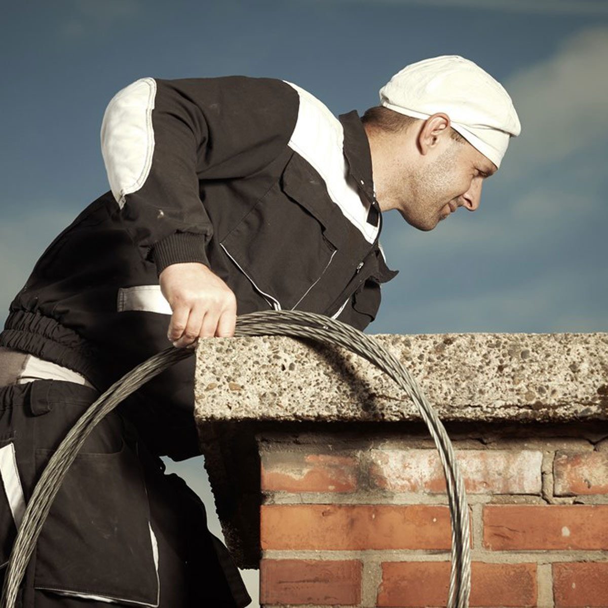 Chimney sweep man in work uniform cleaning chimney on building roof