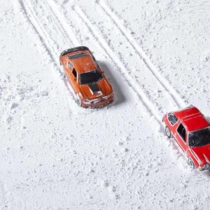 Two toy cars, one orange and one red, leave tire tracks as they move through a snowy surface, suggesting playful racing in a winter setting.
