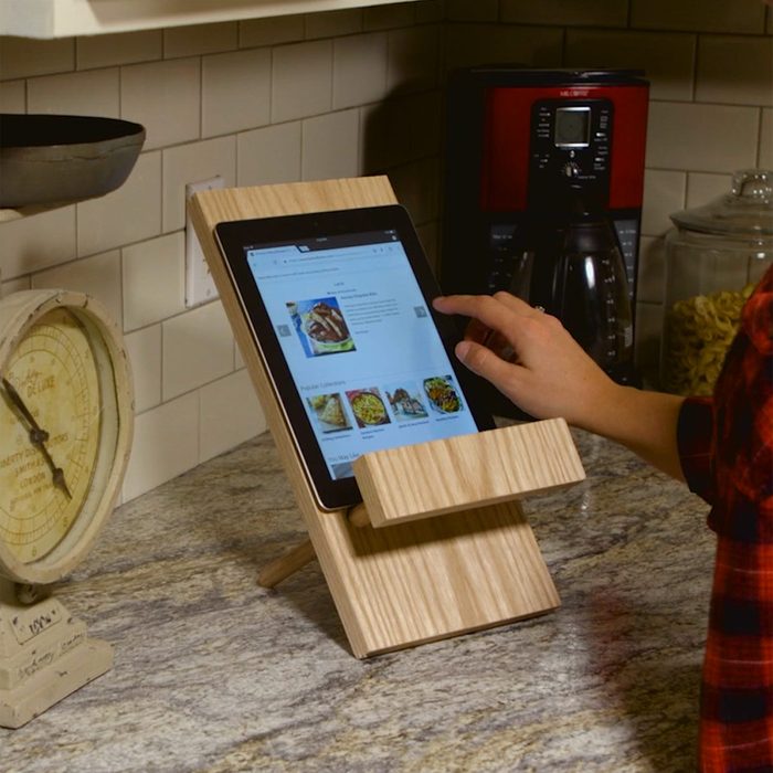 A tablet is positioned upright in a wooden stand, while a hand touches the screen, set on a kitchen countertop near appliances and a scale.