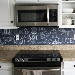 A stainless steel microwave is mounted above a stove, with a chalkboard backsplash featuring inspirational text and decorative flourishes in a kitchen setting.