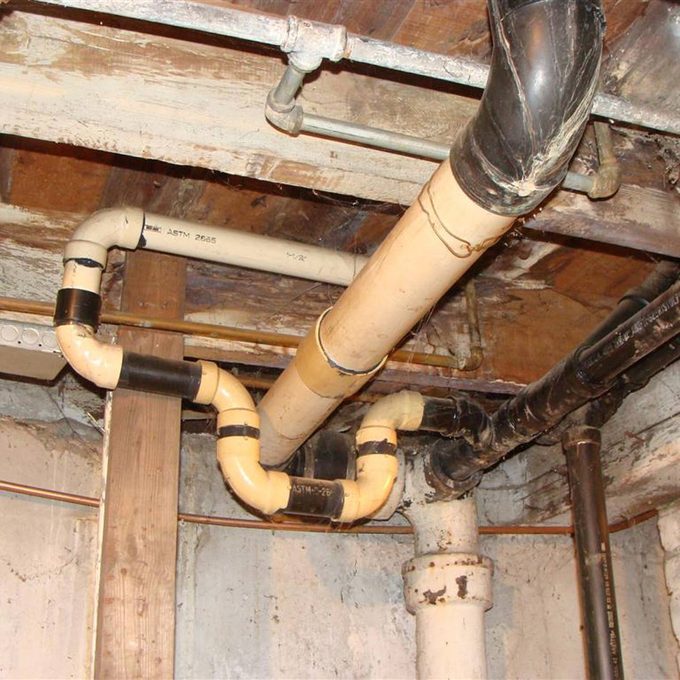 Pipes of various materials and colors are connected in a utility space, with bends and joins near wooden supports, suggesting a plumbing system in a basement.