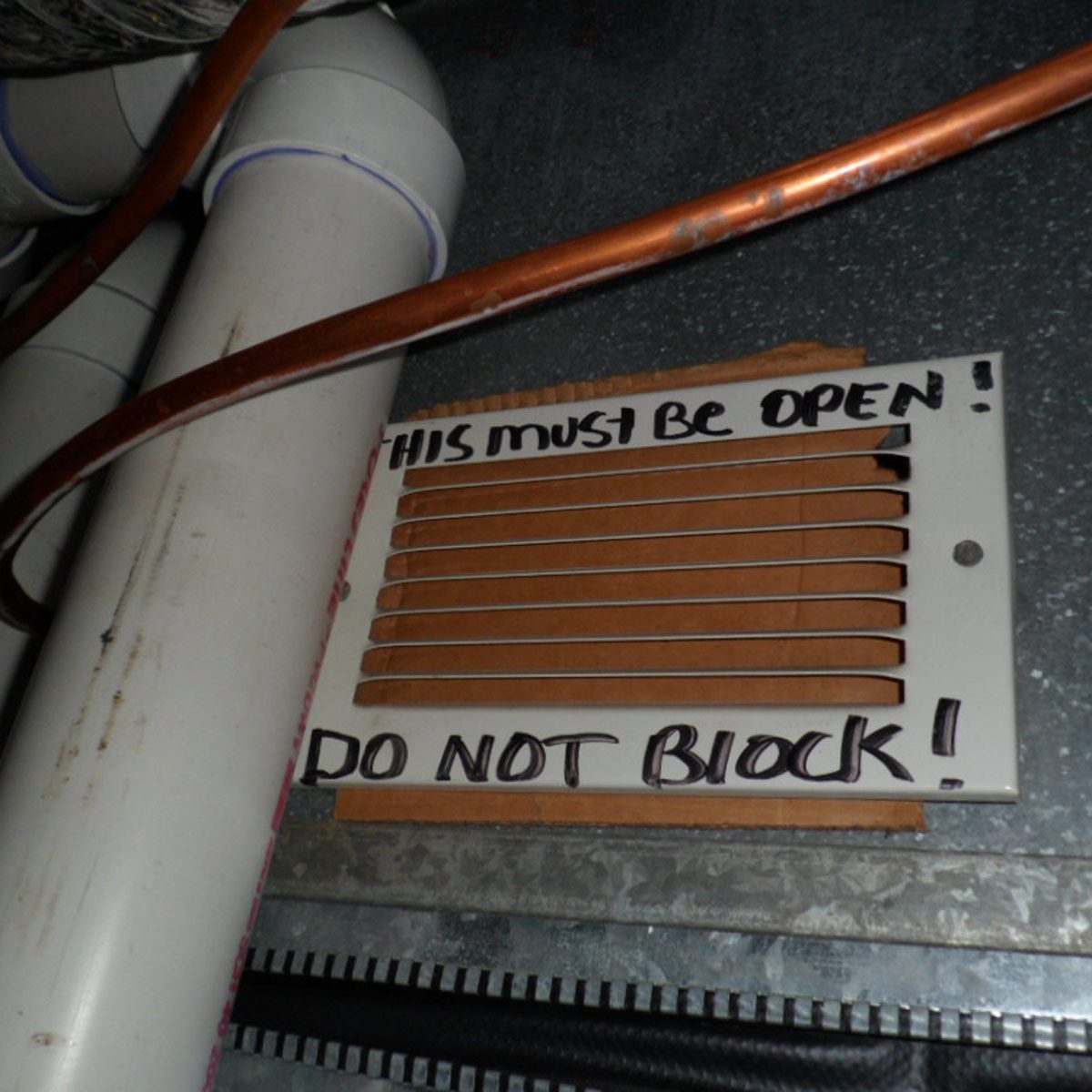 A vent cover with written instructions states, &ldquo;THIS must be OPEN! DO NOT BLOCK!&rdquo; It is surrounded by pipes and an uneven metallic surface.