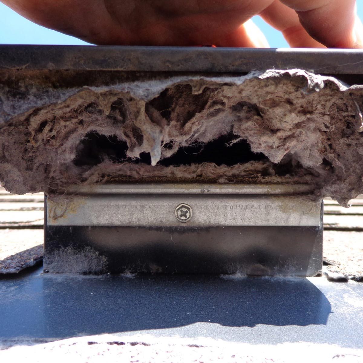 A hand presses against a metal edge revealing a buildup of dust and debris underneath, situated in a sunlit outdoor environment.