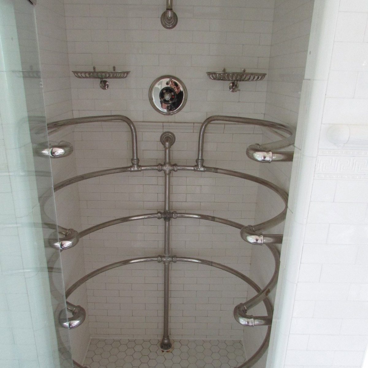 A multi-head shower features curved chrome piping, surrounded by white tiled walls, creating a modern, spacious bathing environment.