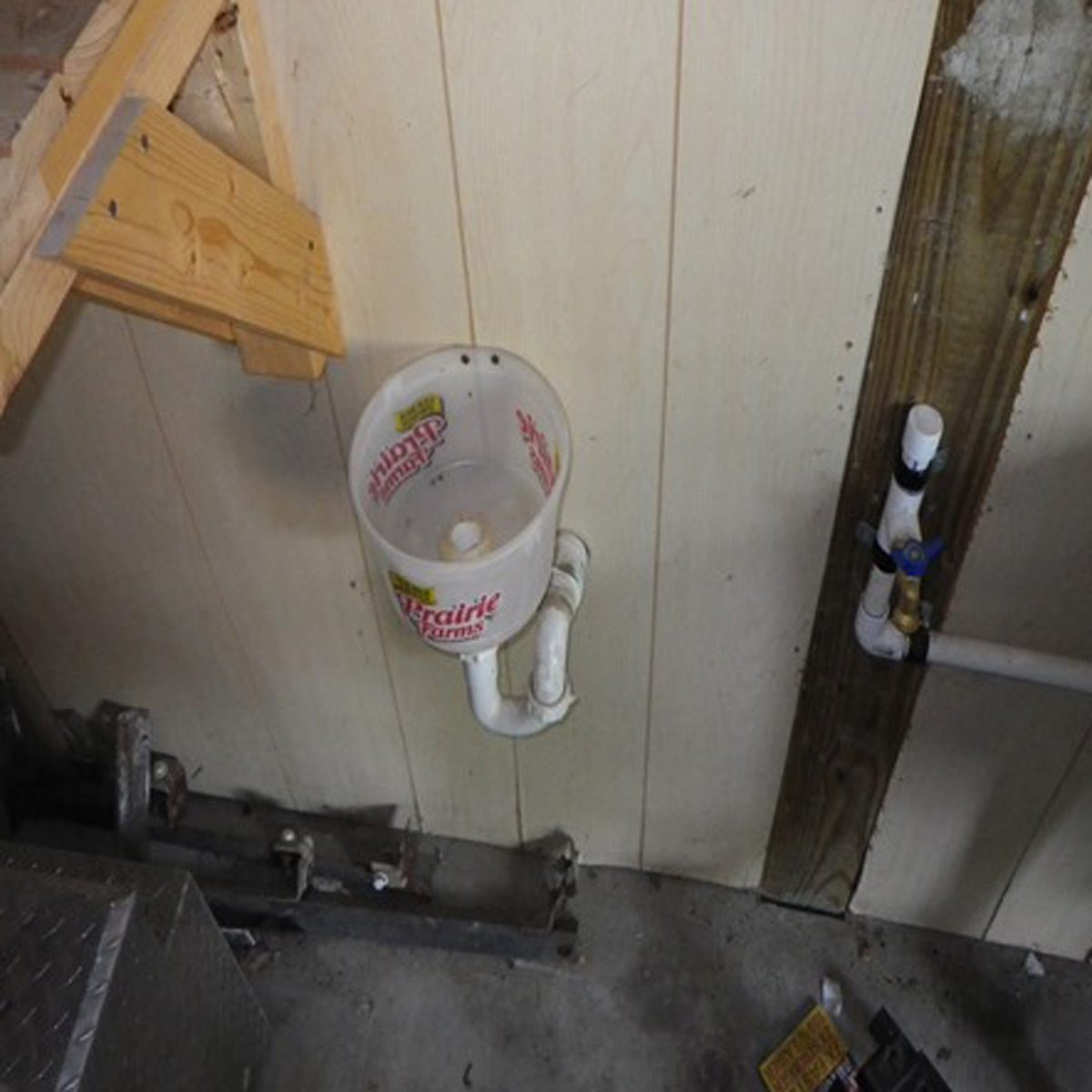 A plastic funnel is mounted on a wall, attached to white pipes in a dimly lit, unfinished area with a concrete floor.