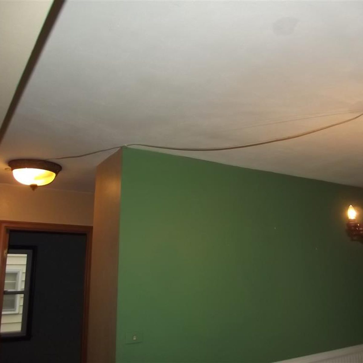 A ceiling light illuminates a room with a green wall. An electrical cord runs along the ceiling towards a light fixture on the opposite wall.