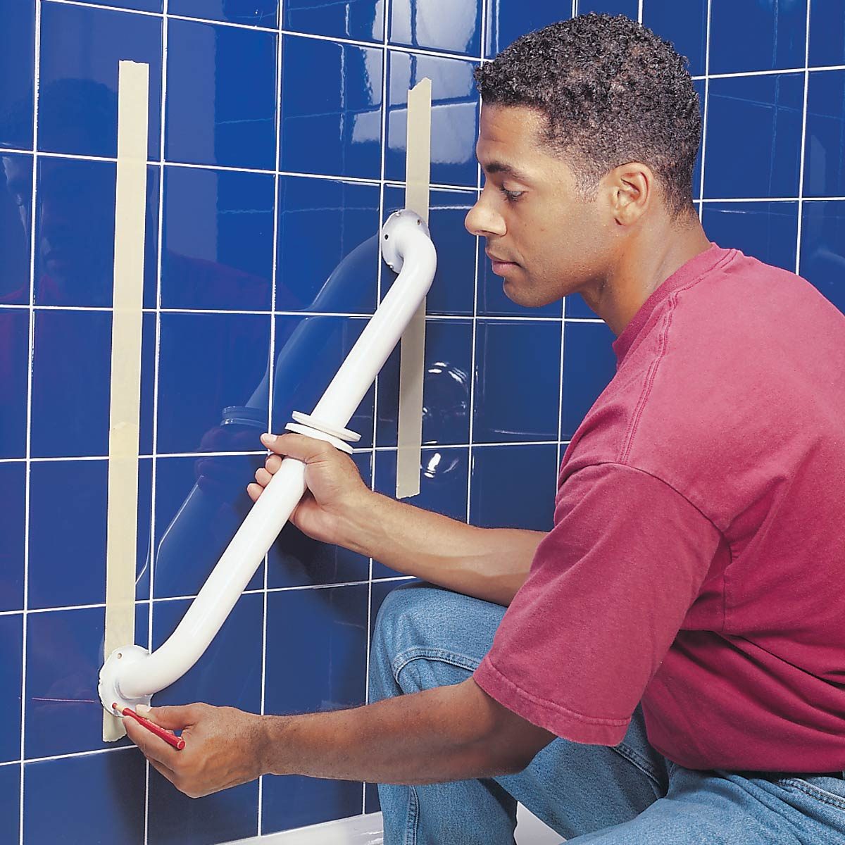 A man positions a grab bar against a blue tiled wall, marking its placement with a red pen while tape aligns the bar for accurate installation.