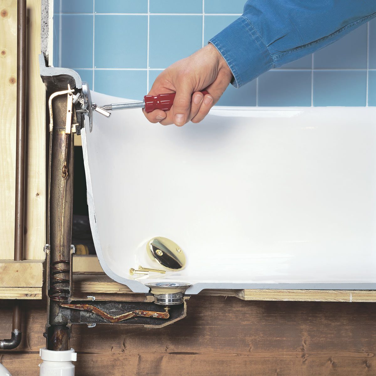 A hand uses a screwdriver to adjust a drain mechanism inside a bathtub, with blue tiled walls and wooden framing visible in the background.