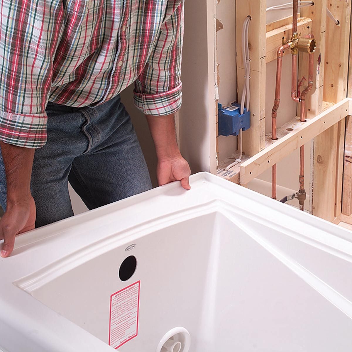 A person is lifting a white bathtub while positioned adjacent to exposed plumbing and framed walls in an unfinished bathroom setting.