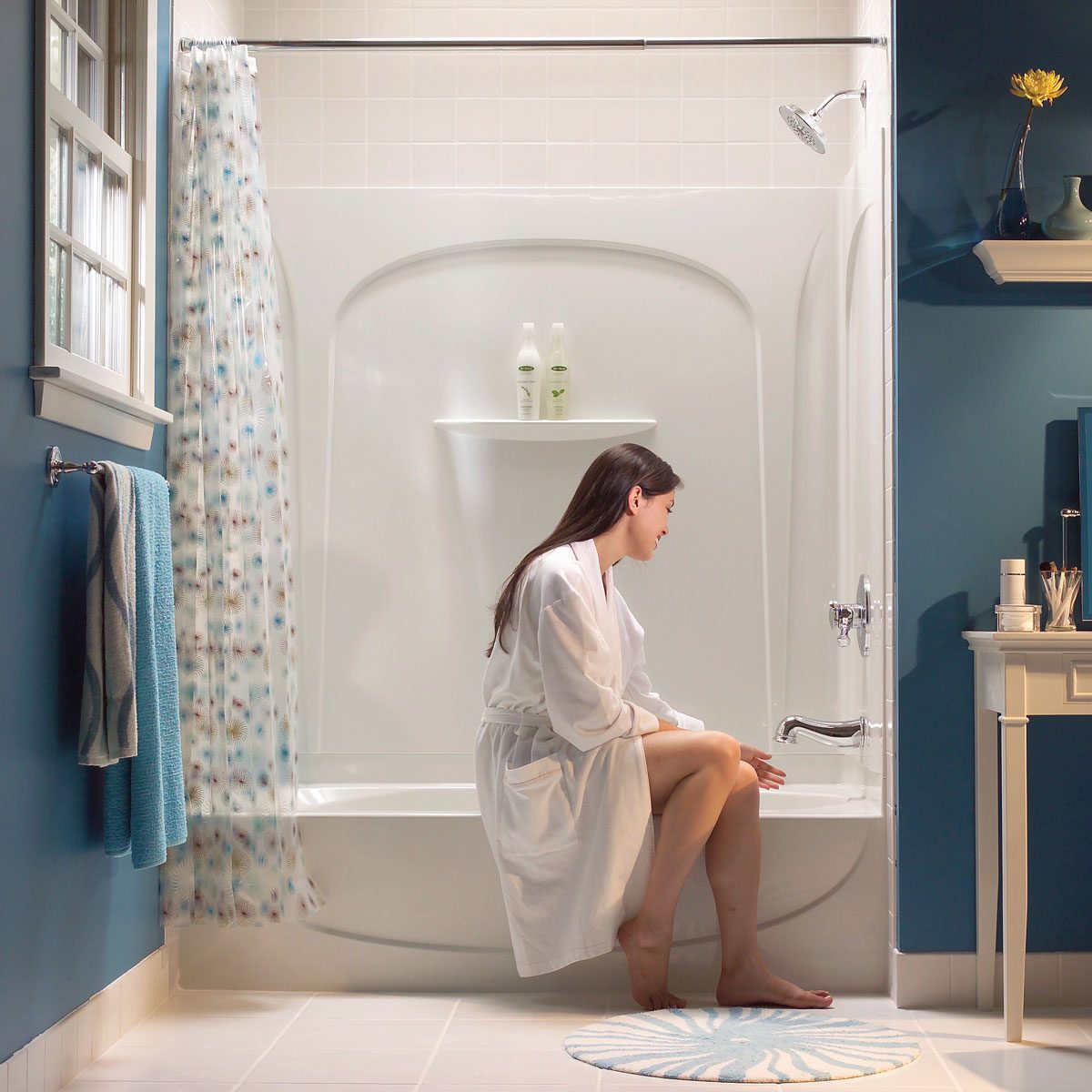 A woman in a white robe sits on the edge of a bathtub, reaching toward the faucet in a stylish bathroom with blue walls.
