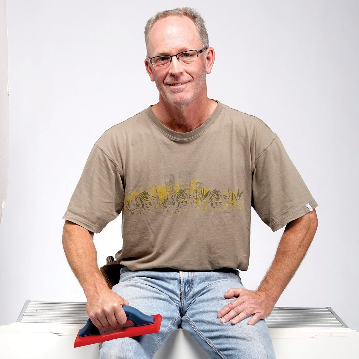 A man sits on a surface, holding a tool with a blue handle and red base, smiling against a simple, light background. Tile Expert Dean Sorem.
