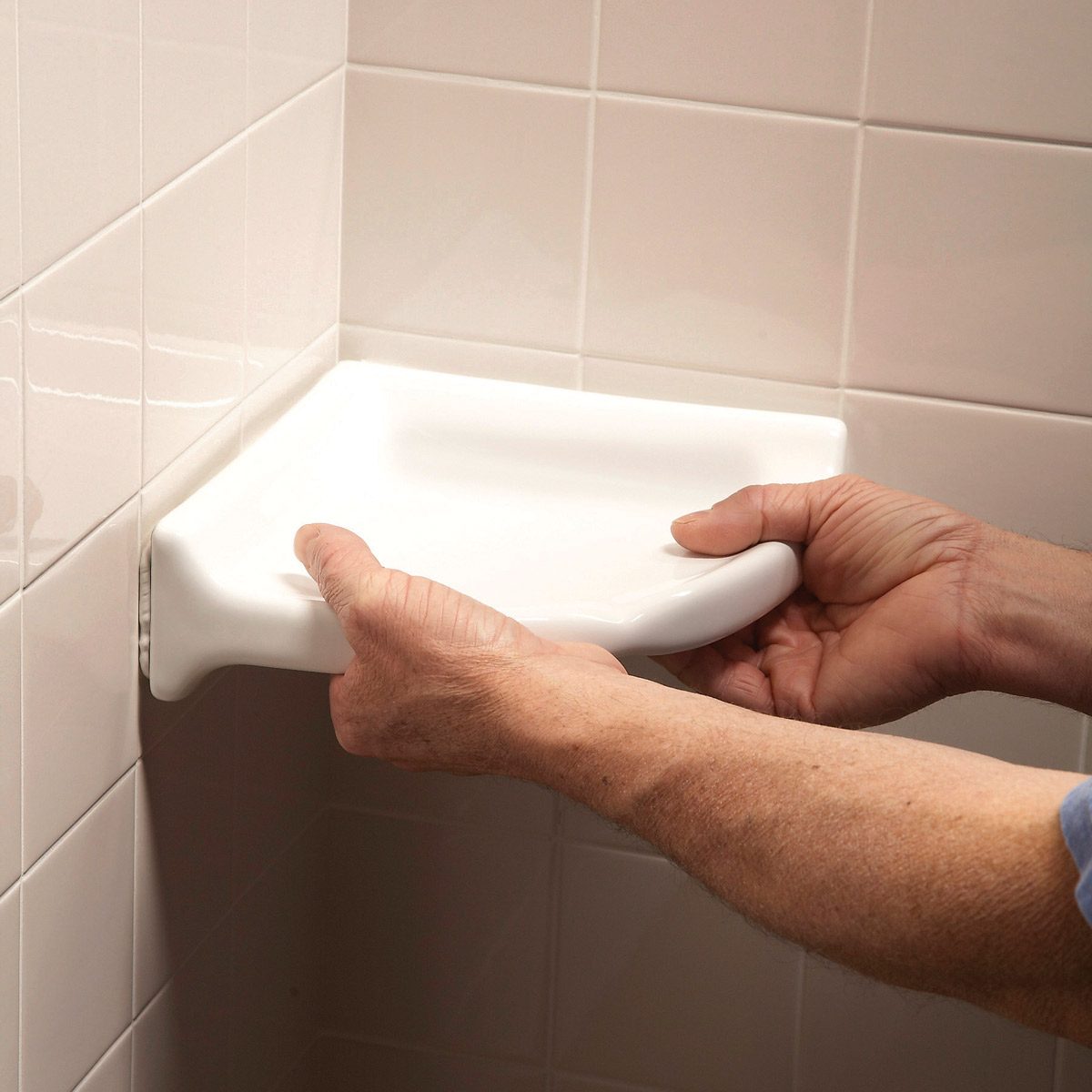 A person is installing a white ceramic corner shelf against tiled walls in a bathroom. Their hands support the shelf, preparing it for secure placement.