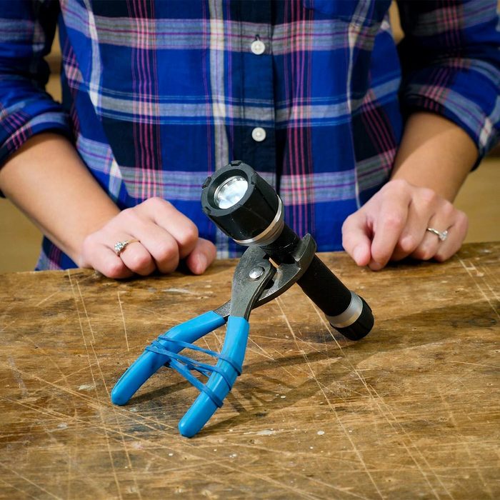 A flashlight is secured between two pliers, held together with rubber bands, resting on a wooden surface while hands approach the setup.