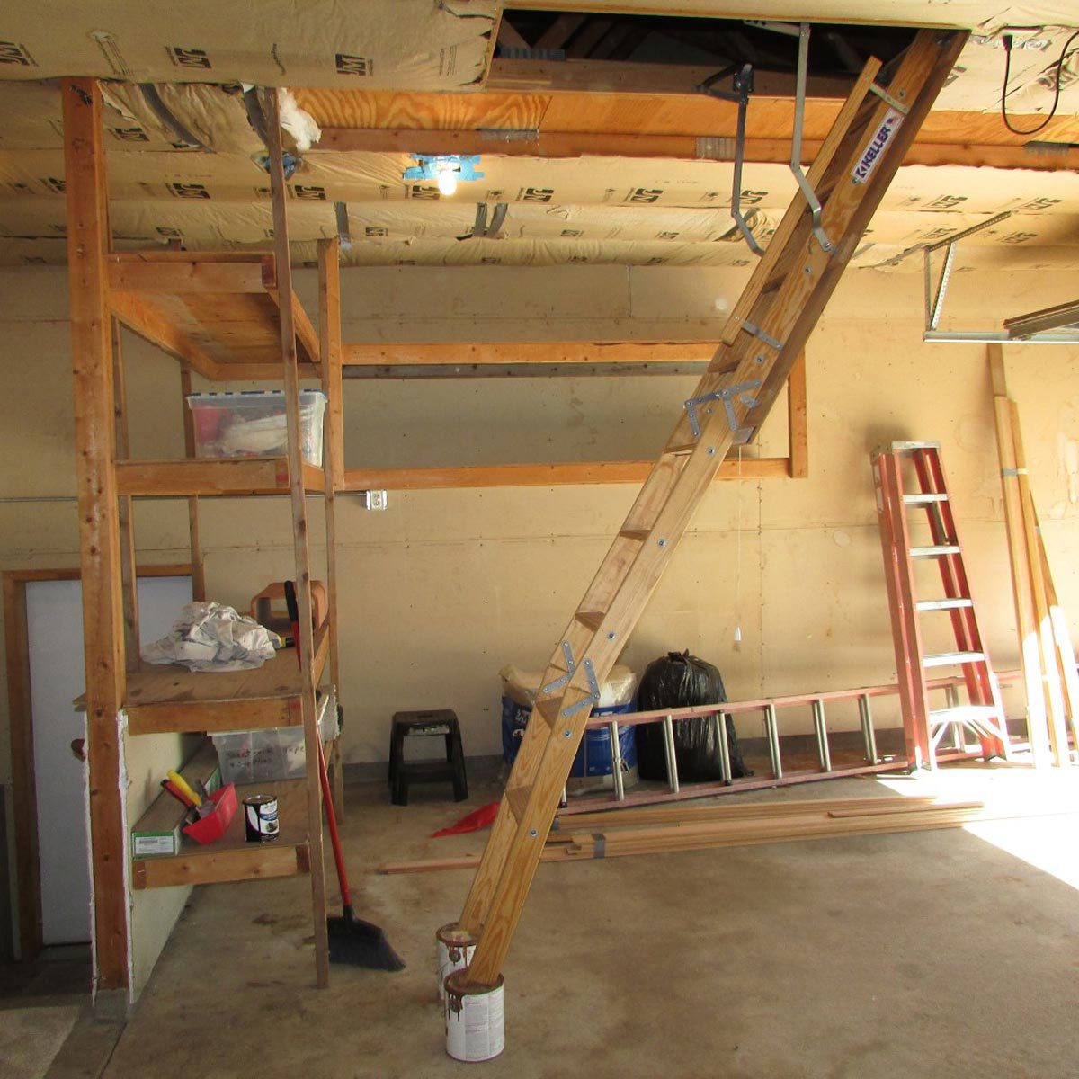 A wooden ladder leans against a structure, connecting upper storage shelves to a garage floor cluttered with tools and materials in a well-lit space.