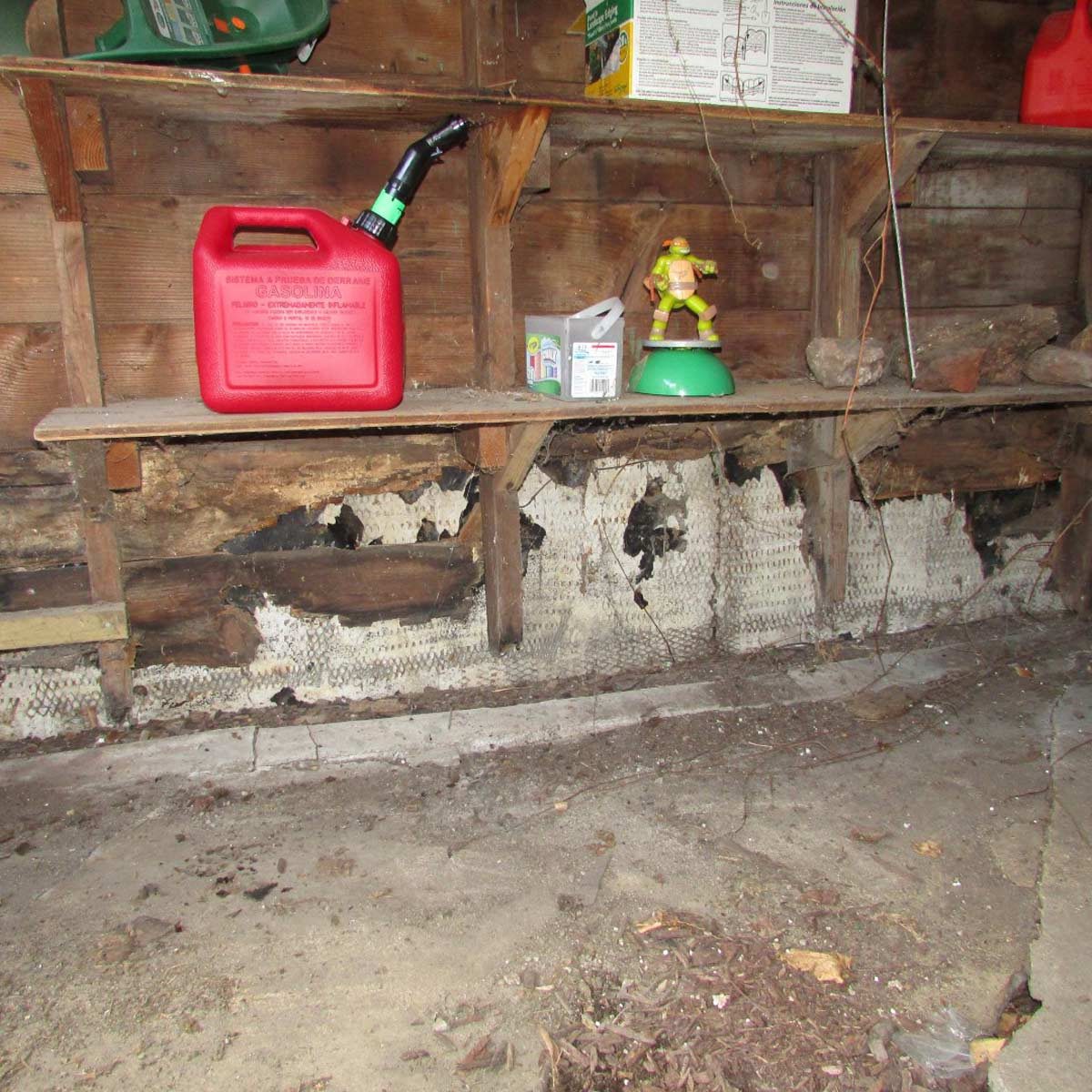 A red gasoline can sits on a wooden shelf in a dusty space, with partially damaged walls and various clutter surrounding it.