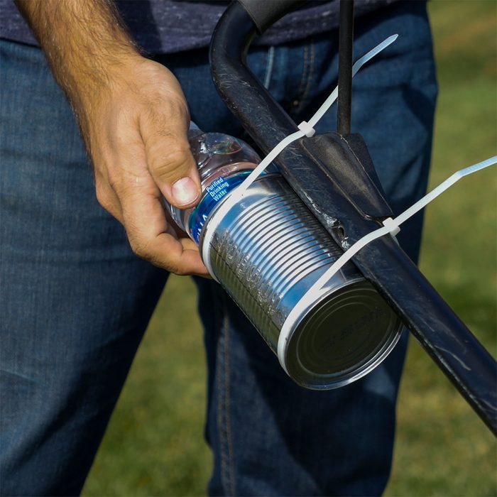 A hand grips a water bottle while securing a can to a black tool with zip ties, set against a grassy outdoor backdrop.