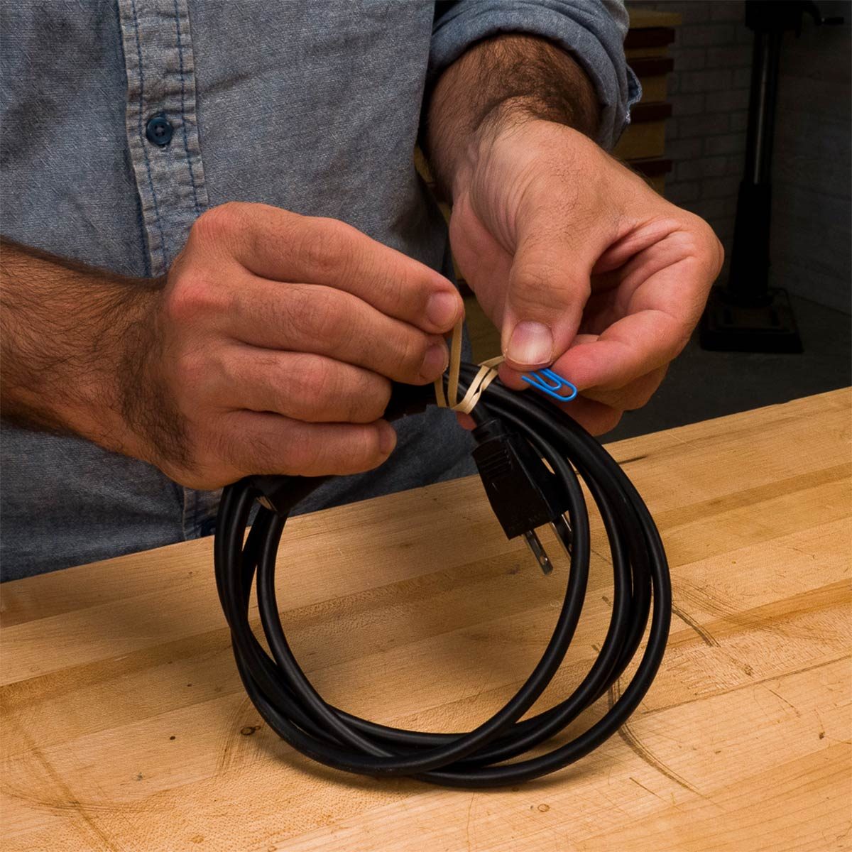 Hands are securing a wrapped electrical cord with a rubber band and paperclip on a wooden work surface in a workshop setting.