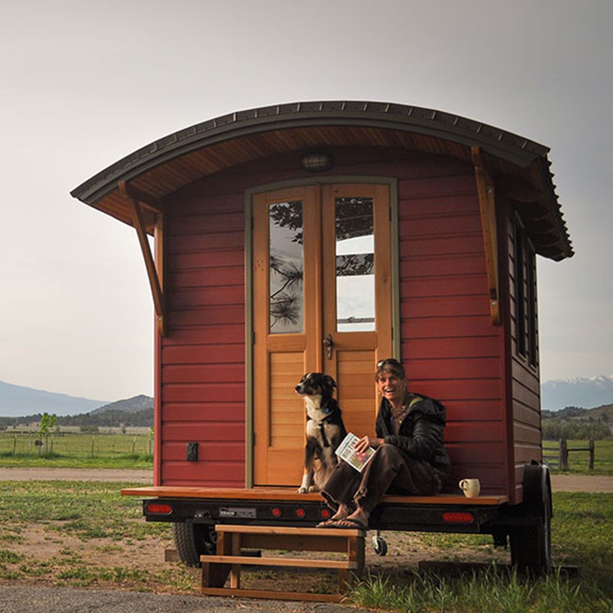 A woman sits on the steps of a red tiny house, reading a book with a dog beside her, surrounded by a grassy landscape and mountains in the distance.
