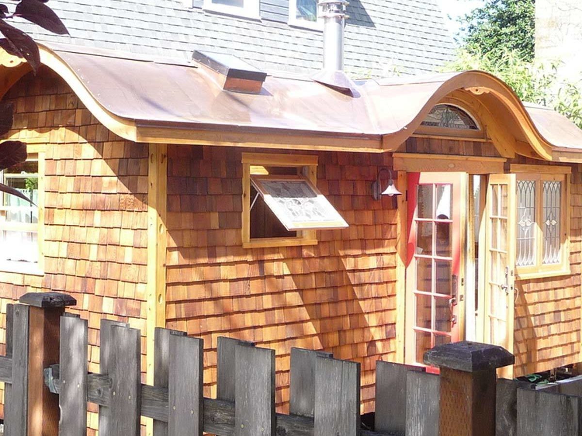 A small wooden house with a curved roof displays a large door and a window, surrounded by a simple wooden fence in a sunny outdoor setting.