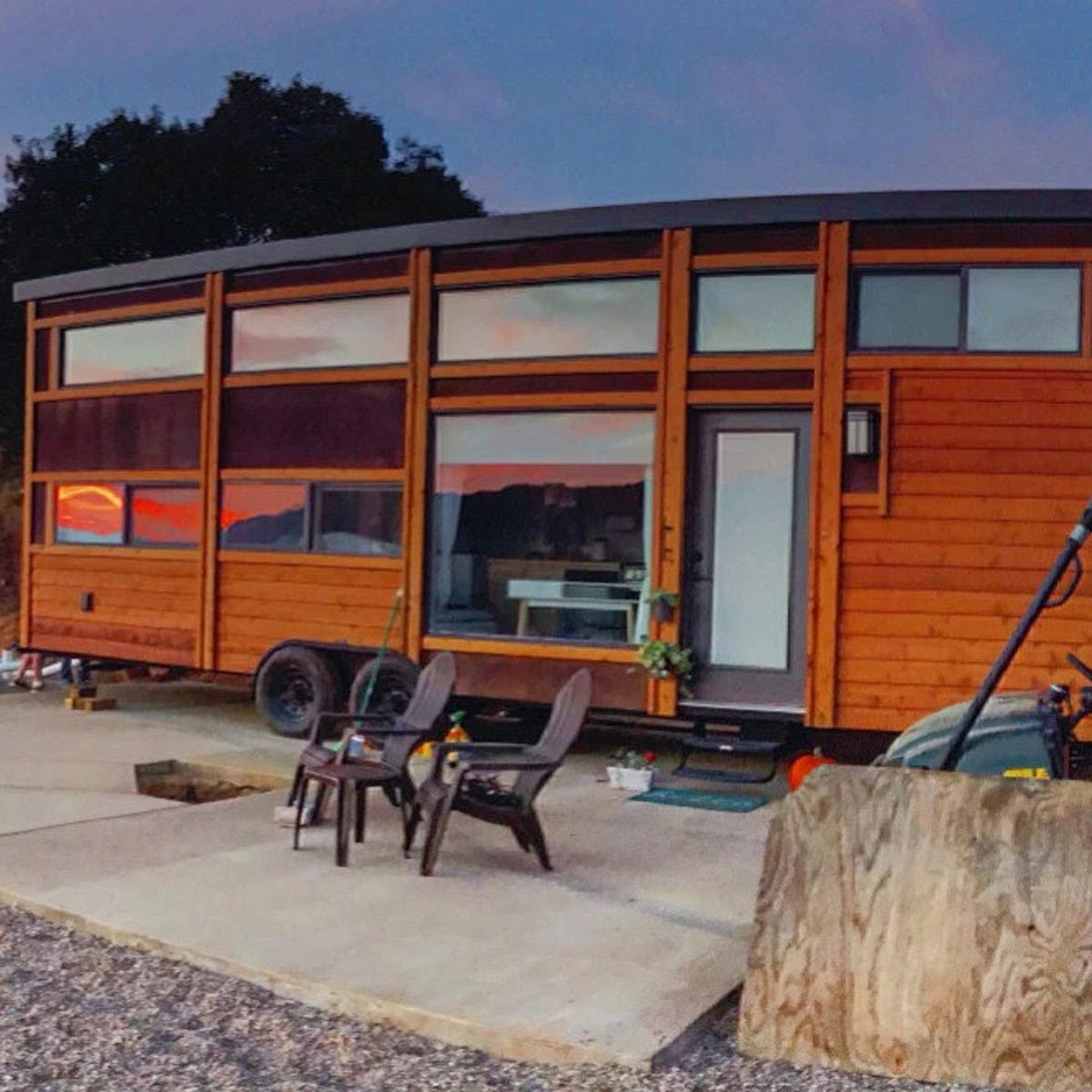 A wooden tiny house sits on a concrete pad, featuring large windows. Two chairs are placed nearby, surrounded by gravel and greenery, under a twilight sky.