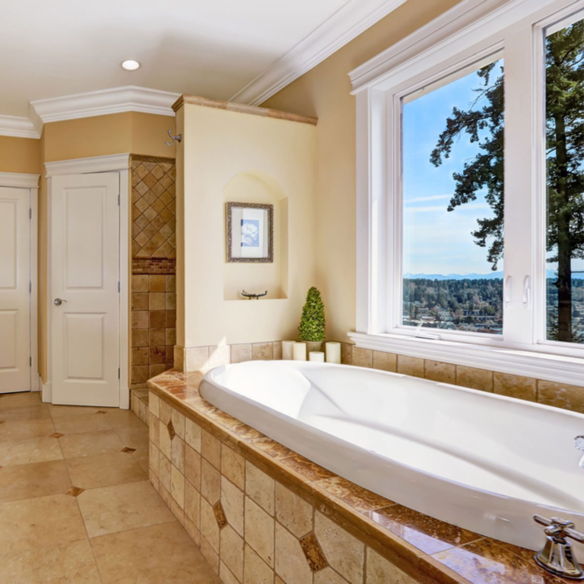 A white bathtub rests on a tiled surface, reflecting light in a bright bathroom with a large window showcasing a scenic view outside.