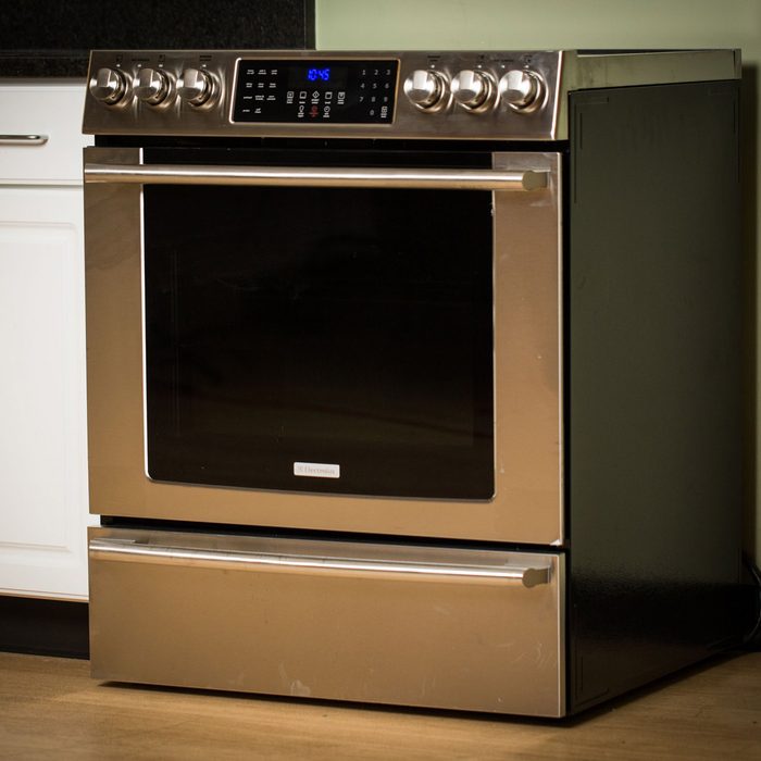 A stainless steel oven stands in a kitchen, displaying a digital clock and knobs on top, with a dark drawer below and a nearby cabinet.