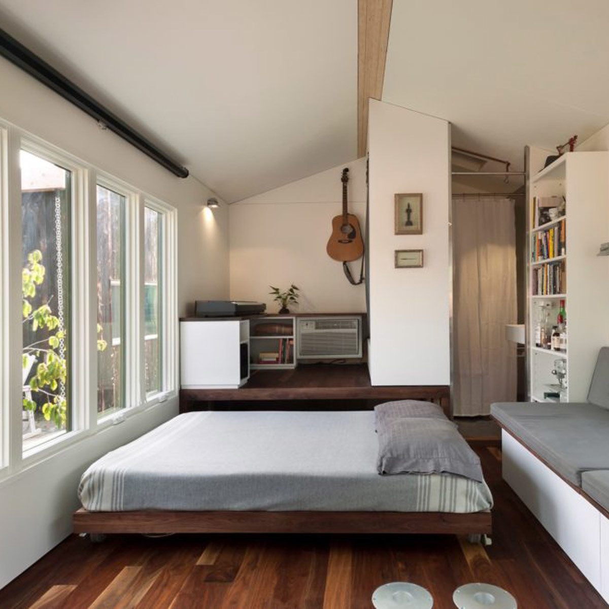 A low-profile bed sits on polished wood flooring in a minimalist room, with a guitar hanging on the wall and sunlight streaming through large windows.