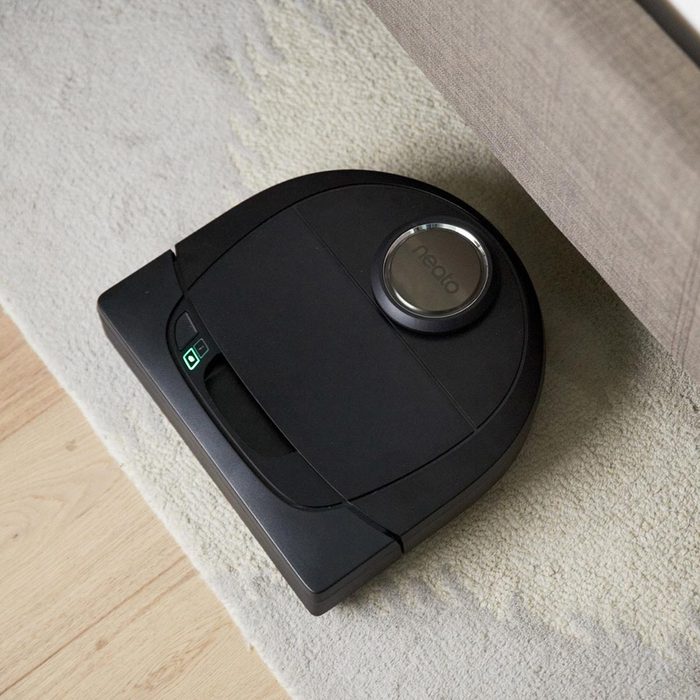 A black robotic vacuum rests on a light carpet, showing a power indicator, positioned under a sofa, as it prepares to clean the surrounding area.