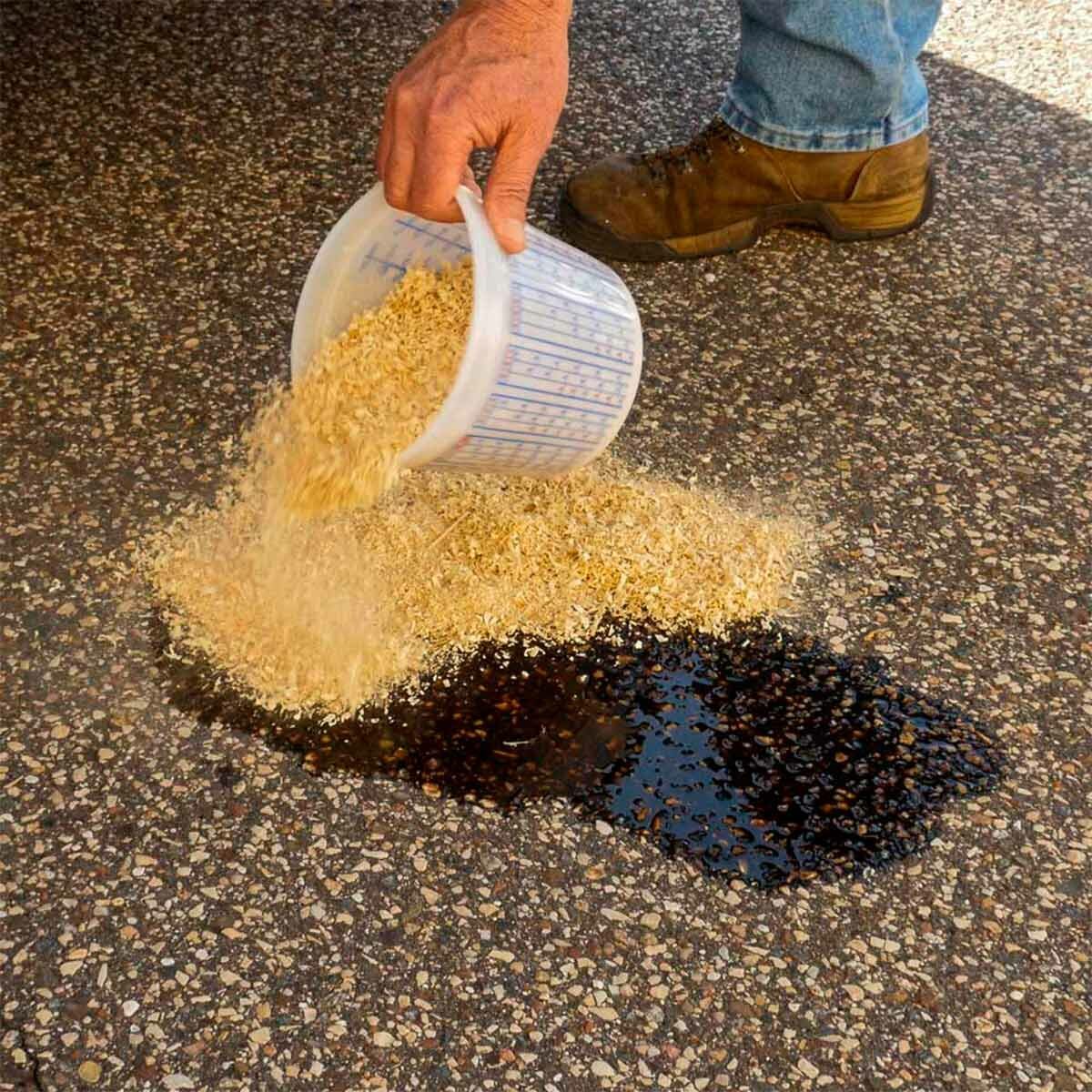 A person pours sawdust onto an oil stain on a textured surface, attempting to absorb the liquid. The environment is outdoors, possibly a driveway.