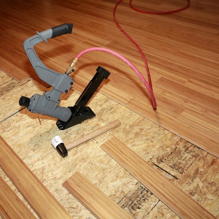 A pneumatic flooring nailer rests on an unfinished wooden surface, ready for use, with planks laid out beside it, while an air hose runs nearby.