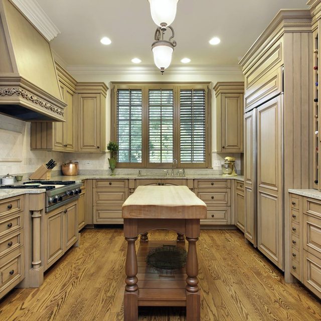 A wooden kitchen island sits center in a well-lit space, surrounded by beige cabinetry and appliances, with sunlight filtering through window blinds.
