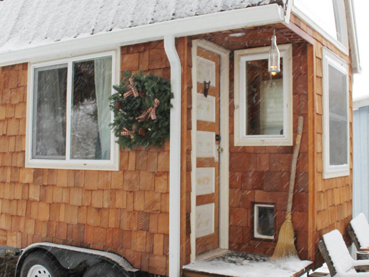 A tiny wooden house is covered in snow, with a wreath on the door and snowflakes falling. The porch has steps and a broom leaned against it.