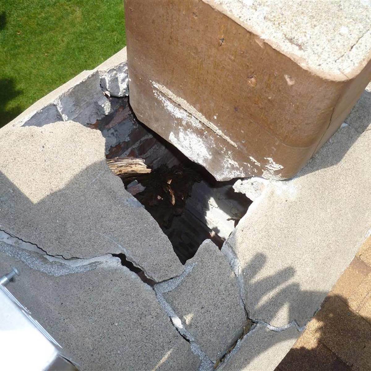 A cracked chimney extends from a rooftop, with a significant gap exposing interior structure and debris, under a clear sky and green grassy surroundings.